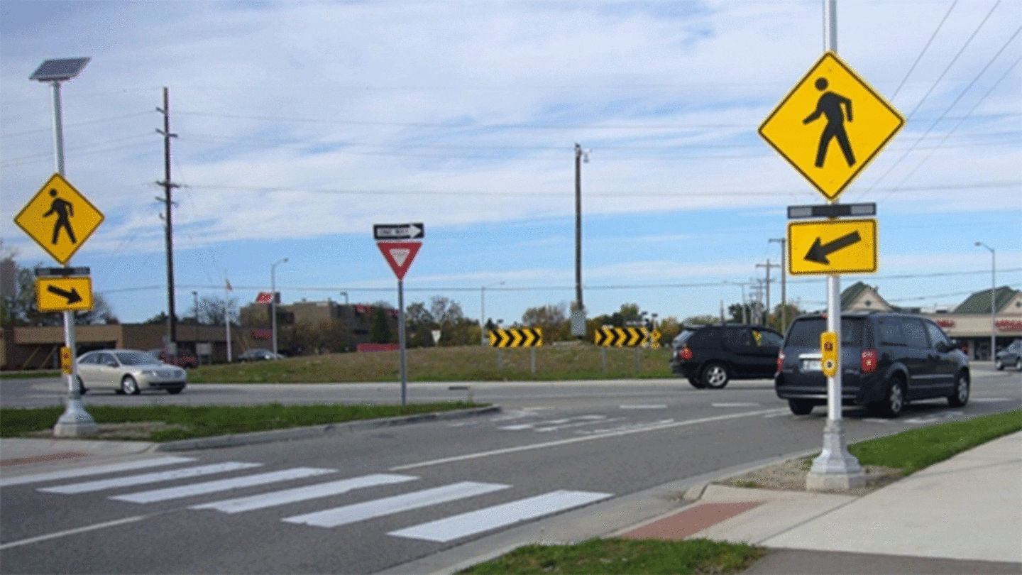 Image of the entry leg of a two-lane roundabout with Rectangular Rapid-Flashing Beacons (RRFBs) installed on both sides of the crosswalk. The RRFB are rectangular flashing yellow lights mounted inbetween a pedestrian sign and an arrow sign pointing to the crosswalk.