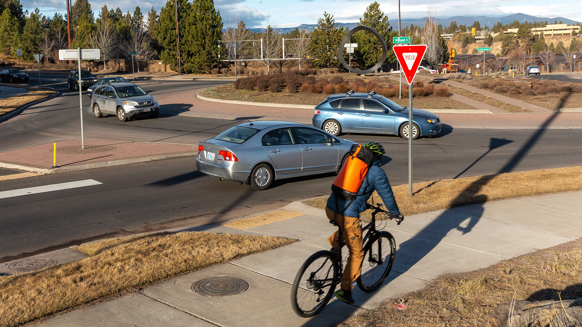 Image of an entering leg of a single-lane roundabout. A bicyclist is seen approaching the roundabout on the sidewalk, while several vehicles are seen traveling through the roundabout. The roundabout corners have the PROWAG-required edge treatment in the form of landscaping.