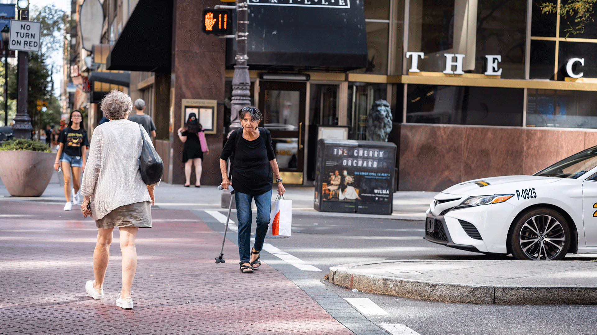 Image of a woman with a walking cane crossing at an urban intersection with a pedestrian countdown signal along with other pedestrians. A vehicle is seen yielding to the pedestrians at this signalized intersection crossing.