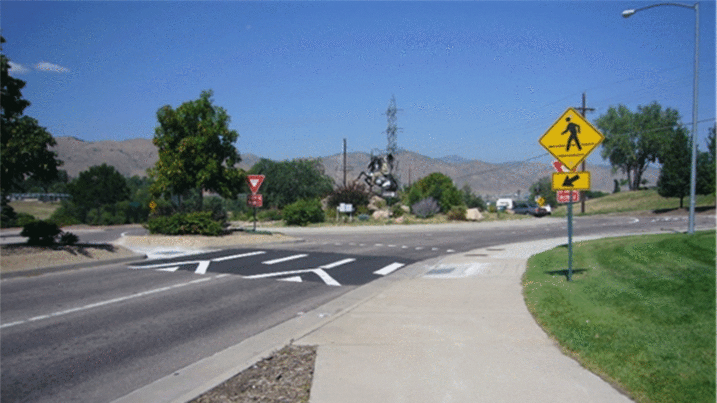 Image of the entry leg of a two-lane roundabout with a raised pedestrian crossing installed. The raised crossing is elevated by 3-5 inches relative to the vehicular travel lane, resulting in drivers having to slow down as they approach and traverse the crosswalk.